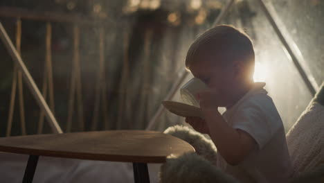 kid drinks tea at small table at glamping. little boy takes sip of drink from cup on pillow in evening camp. child with tasty beverage on terrace