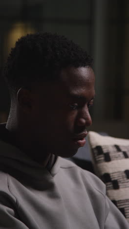 vertical video close up of unhappy and depressed young man sitting on sofa at home looking anxious and worried resting head in hands