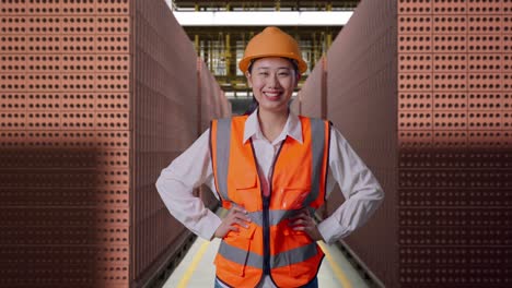 Asian Female Engineer Wearing Safety Helmet Smiling To Camera While Standing With Arms Akimbo With Red Brick Packed in Stacks Are Stored