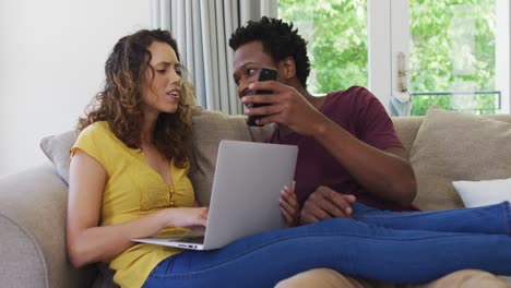 feliz pareja biracial sentada en el sofá con computadora portátil y teléfono inteligente y riendo