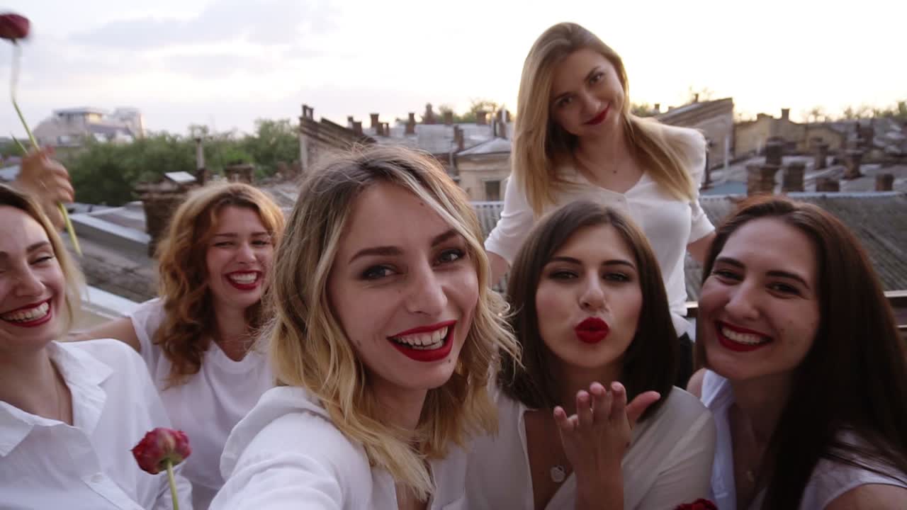 젊은 여성들의 그룹은 테라스에서 놀고, 즐기고, 웃고 있습니다. 카메라를 위해 포즈를 잡고, 셀카를 찍고 있습니다. 모두  ⁇ 색과  ⁇ 은 립스틱을 입고 있습니다. 배경에 아름다운 오래된 도시 전망