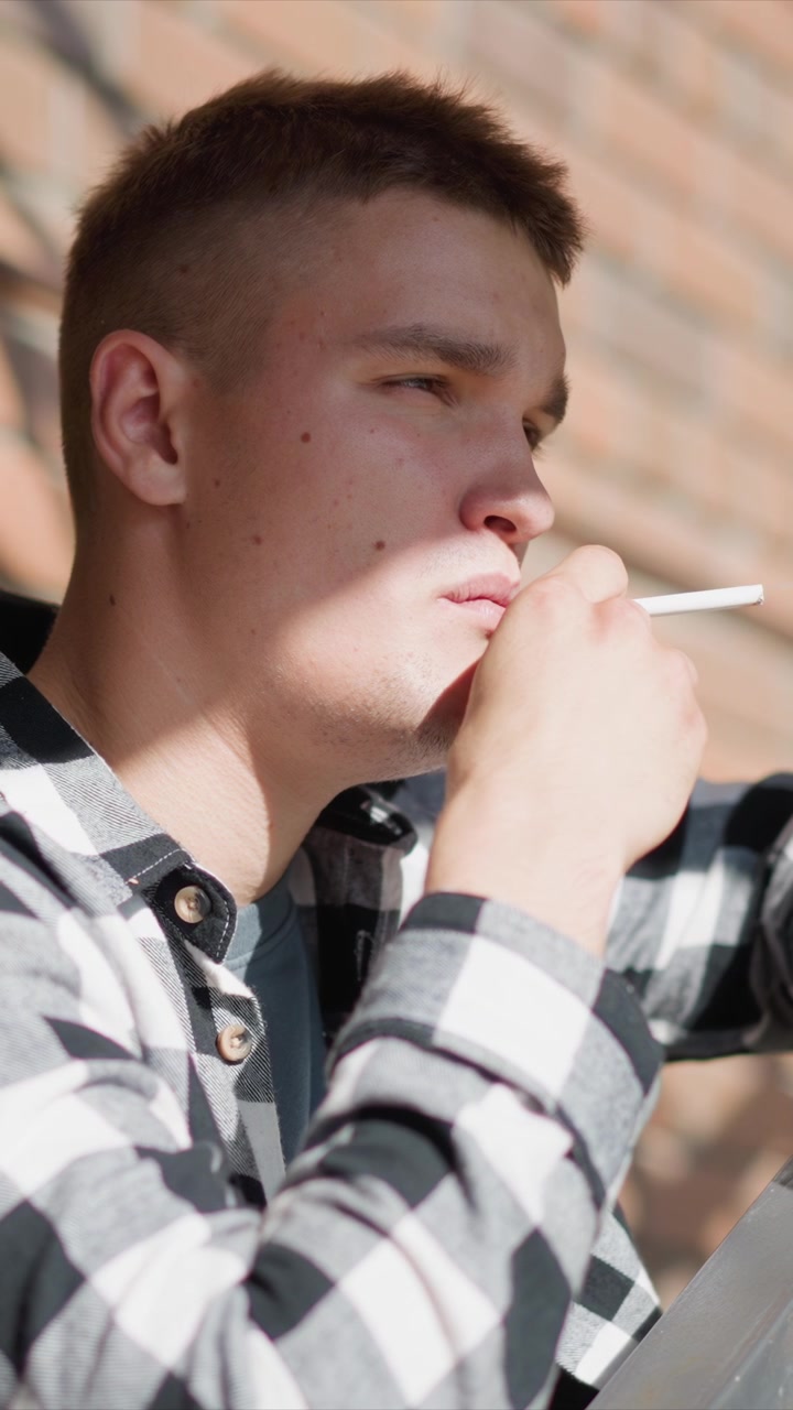 Primo piano di giovane uomo che tiene una sigaretta in mano ed espira fumo. L'uomo indossa una camicia a quadri bianchi e neri con bottoni visibili su sfondo di muro di mattoni. La luce naturale soffusa crea