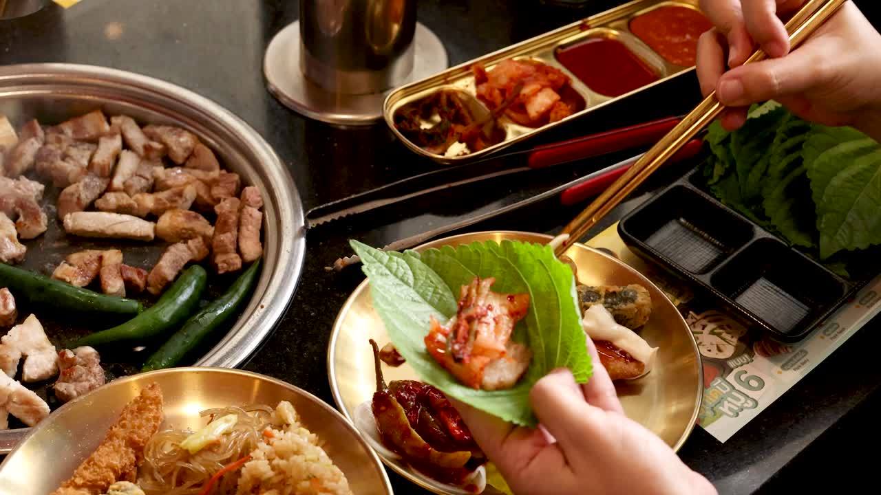 Hand prepares lettuce wrap with grilled pork, kimchi, vegetables at Korean BBQ table, warm lighting