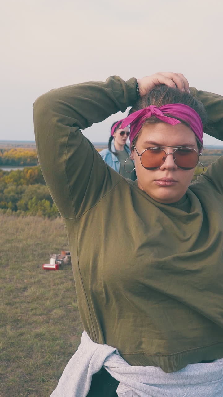 pretty overweight girl fixes hair by friends resting at campsite on green bank against calm river at sunset