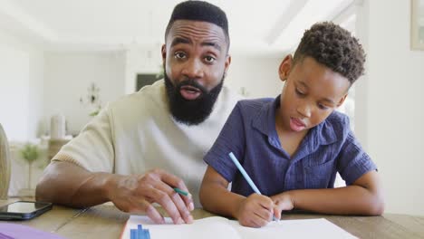 Glücklicher-Afroamerikanischer-Vater-Und-Sohn-Sitzen-Am-Tisch-Und-Machen-Gemeinsam-Hausaufgaben,-In-Zeitlupe