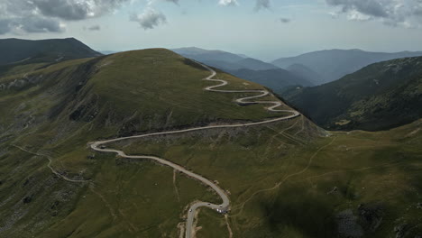 Aerial-view-of-winding-roads-through-the-lush-green-hills-of-Transalpina-in-Romania,-with-mountain-ranges-in-the-distance