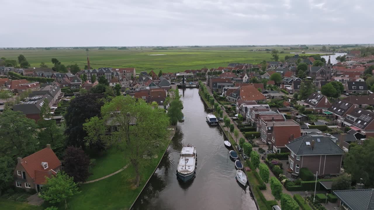 A Luxemotor ship sails along a narrow canal through a Dutch town surrounded by greenery in the Netherlands. Zoom out