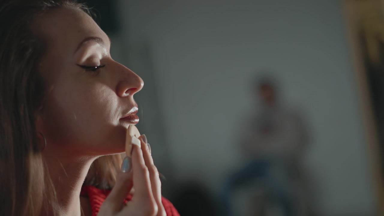 Giovane donna con un maglione rosso di maglia pulisce delicatamente la palpebra inferiore e la guancia con un dischetto di cotone, facendo movimenti delicati verso l'alto mentre rimuove i residui di trucco, con gli occhi chiusi, espressione calma, al chiuso.