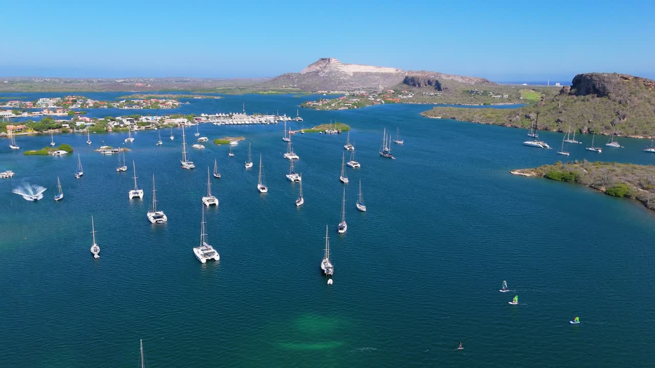 Aerial panoramic orbit of yachts and sailboats anchored in clear blue waters, surrounded by scenic coastline and distant hills