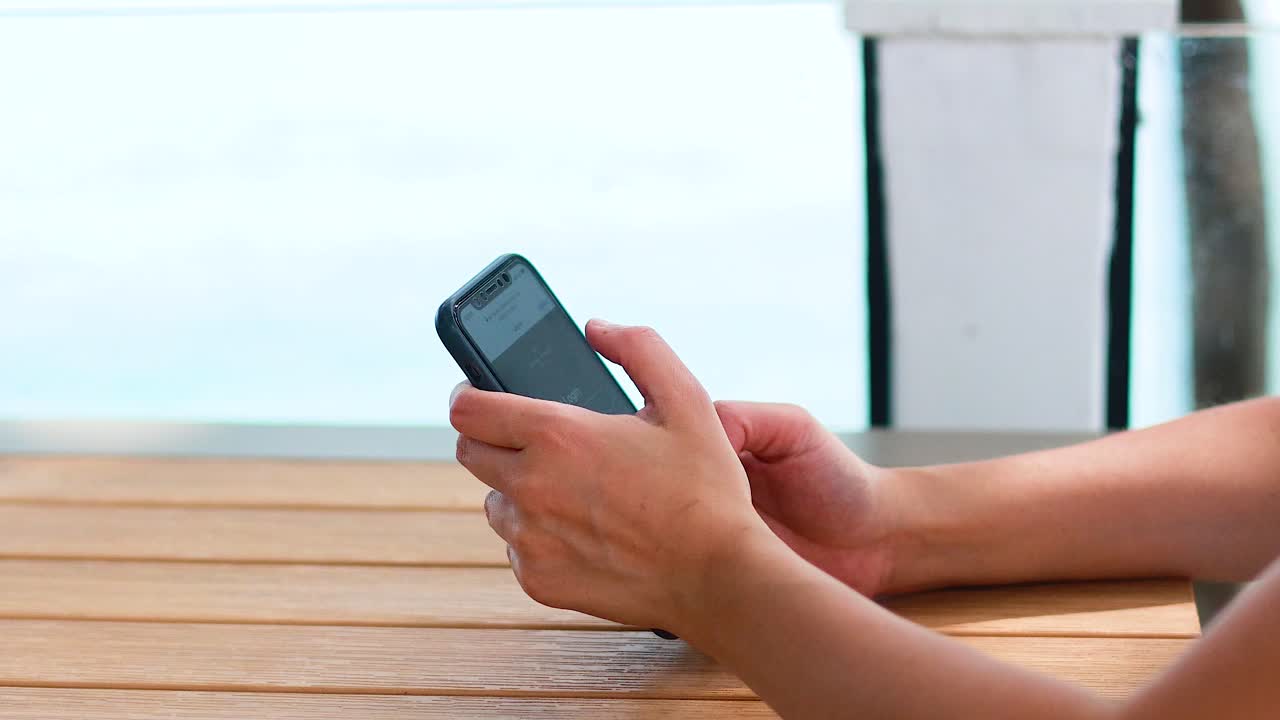 A person interacts with a smartphone on a wooden table near the beach in Phuket, Thailand. Bright, natural lighting