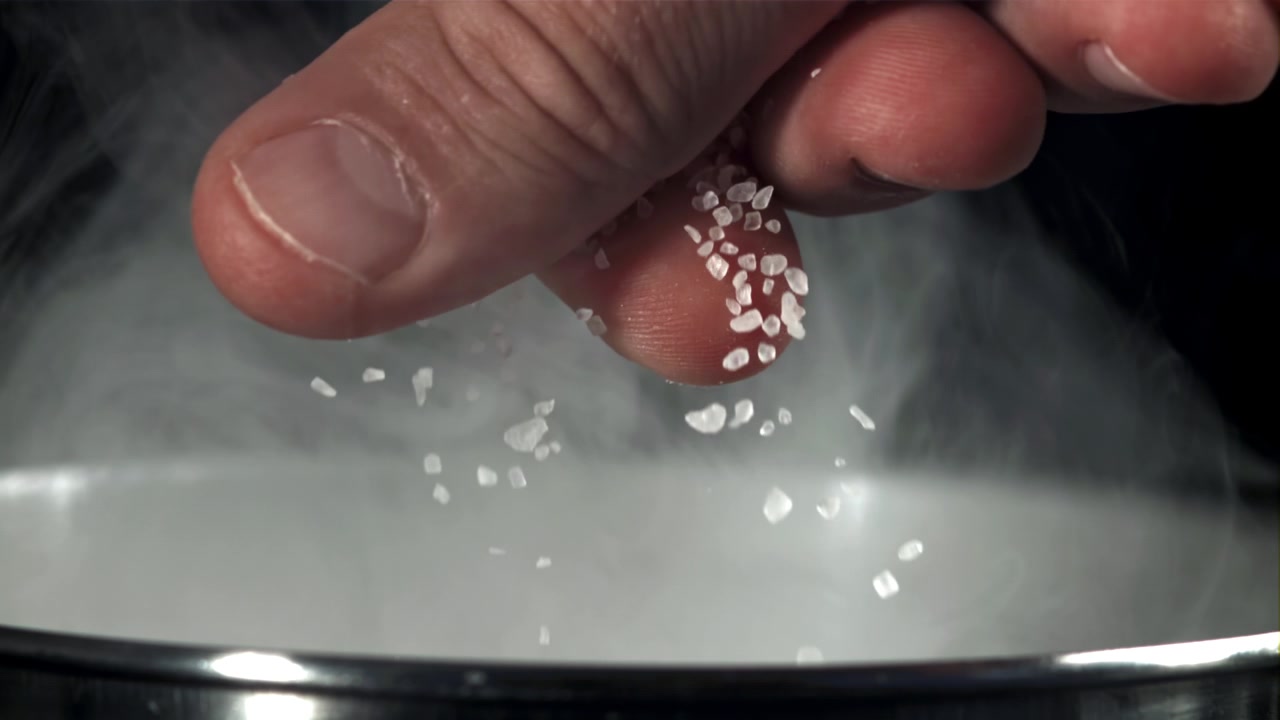 Nærbillede af hånd, der drysser salt ned i en dampende gryde i slowmotion
