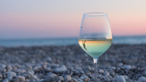 glass of white wine reflecting silhouette of woman walking on pebbles beach at twilight