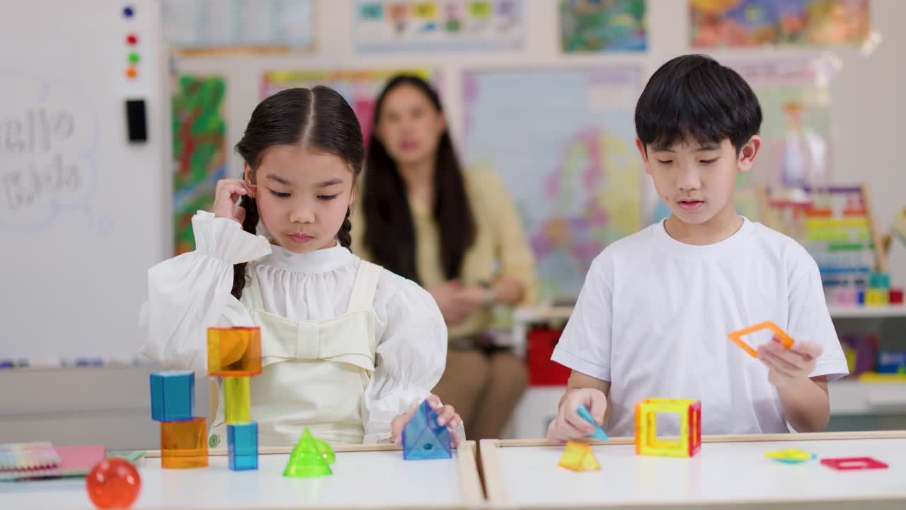 Two children construct colorful geometric shapes with blocks at a classroom table, teacher observing behind