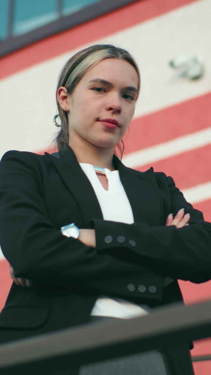 Lady in black blazer folds hands confidently while standing in front of organization building with red and white stripes and visible security camera, projecting strength