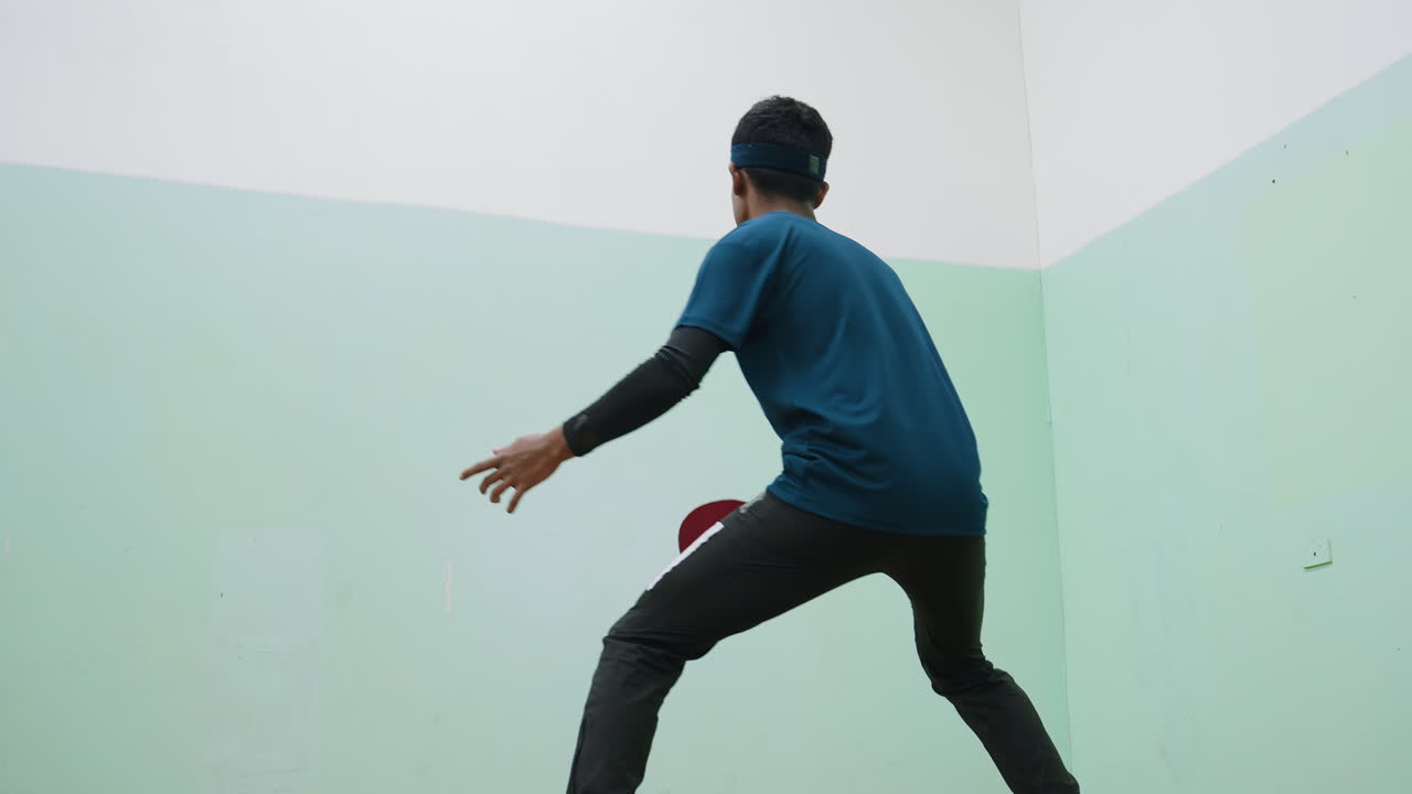 Young man wearing headband and sportswear practices tennis against wall indoors, standing in active stance holding paddle, focusing on ball movement to improve skills