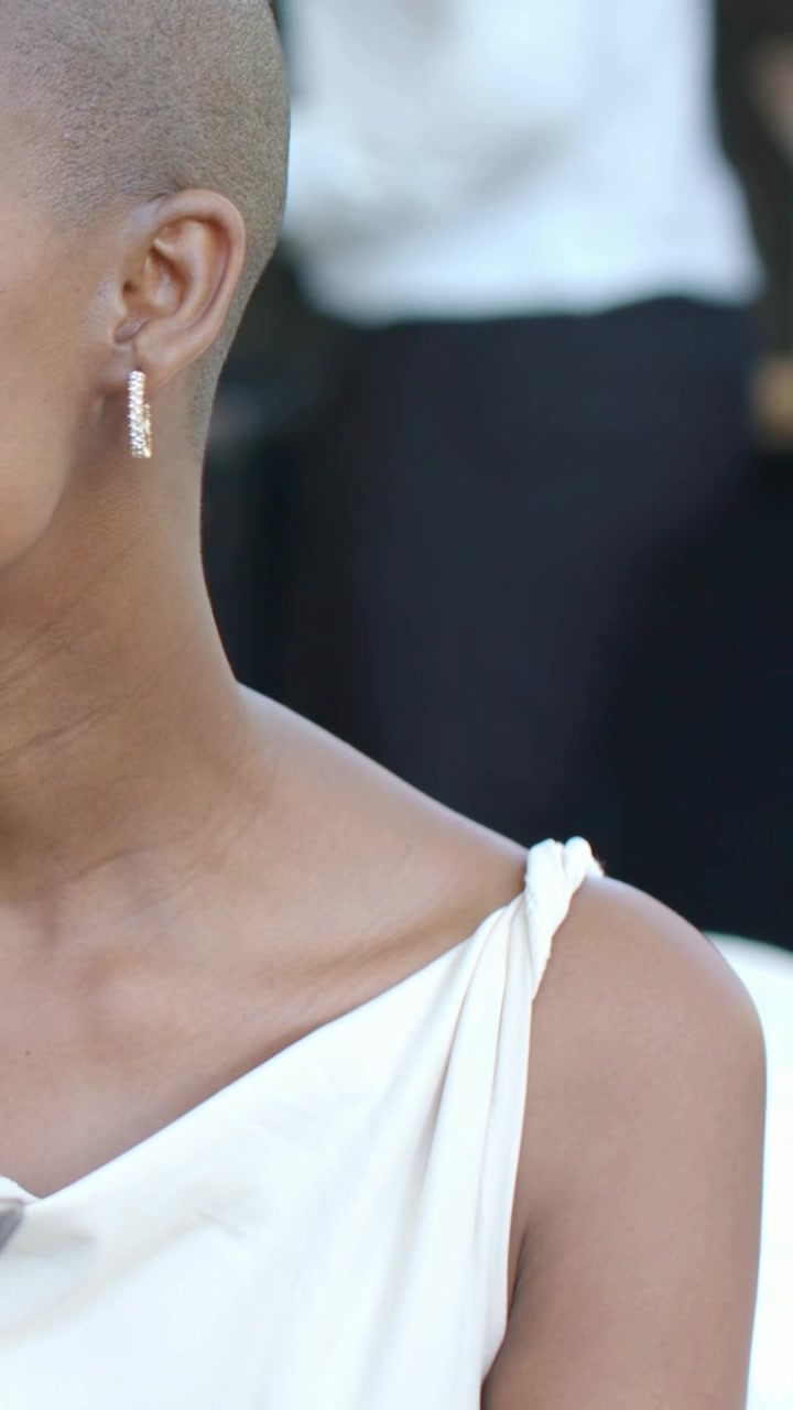 Video verticale: Una donna si volta e guarda a sinistra a un gala, mentre il personale si muove; indossa orecchini a cerchio e un vestito bianco. Spazio per il testo.