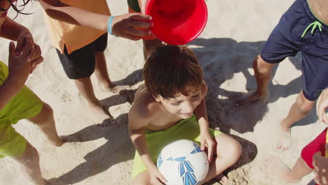 boy pouring water on sitting on beach friends head from bucket