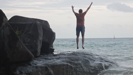 Back-View-Of-A-Man-Jumping-On-A-Rock-Facing-The-Sea-At-Dam-Trau-Beach-In-Con-Dao-island,-Vietnam