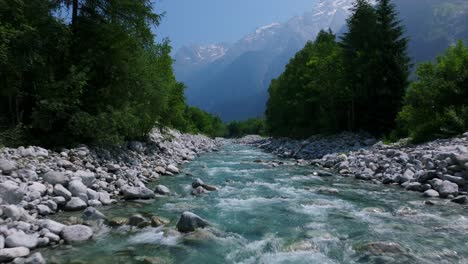 drone flying over pristine alpine river, rocky riverbed, green forest, and majestic mountains in background, switzerland. aerial forward at low altitude