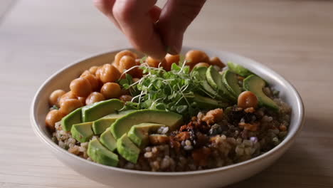 hand garnishing a healthy vegan bowl with microgreens, avocado, and chickpeas