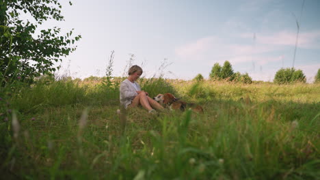 woman seated on ground with her dogs lying close by, enjoying nature in grassy field under sunny sky, dogs gaze attentively with tongue out while owner focuses on something unseen