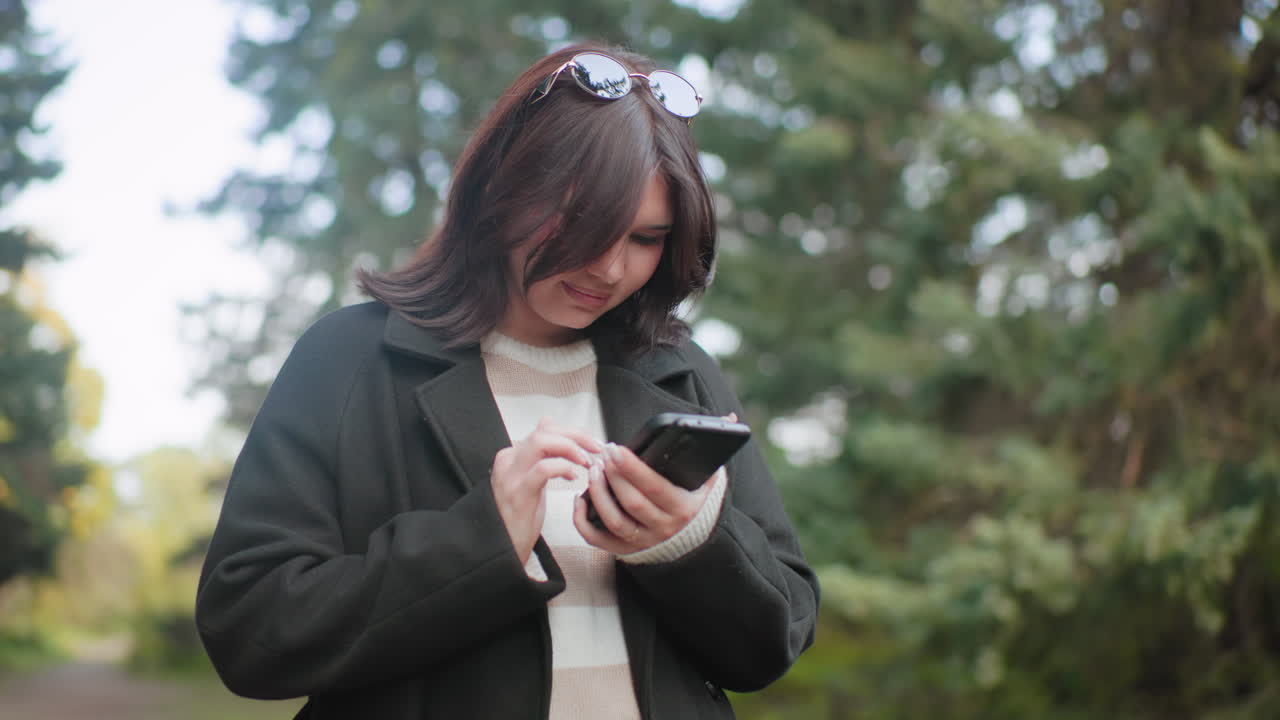 수목원에서 두 개의 휴대폰 화면을 넘기며 서 있는 자연을 사랑하는 사람, 부드러운 보케 나뭇잎, 긴 코트, 머리에 선글라스를 얹고, 집중하는 미소, 차분한 가을 분위기, 상록수 사이에서 느린 야외의 순간