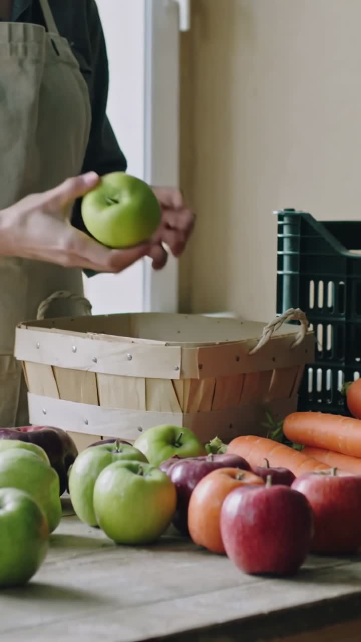 Video vertical: Mujer con delantal extendiendo la mano izquierda recogiendo manzanas verdes en la mesa, colocándolas en una cesta para empacar.