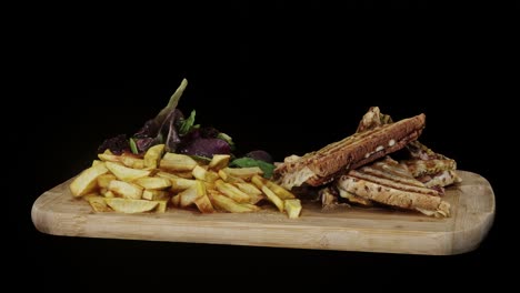 vista lateral de un sándwich a la parrilla en un plato de madera giratorio con papas fritas en el lado, ensalada verde y mayonesa, fondo negro