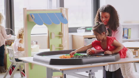 Young-schoolgirl-standing-at-a-sensory-play-table-with-female-teacher-in-an-infant-school-classroom