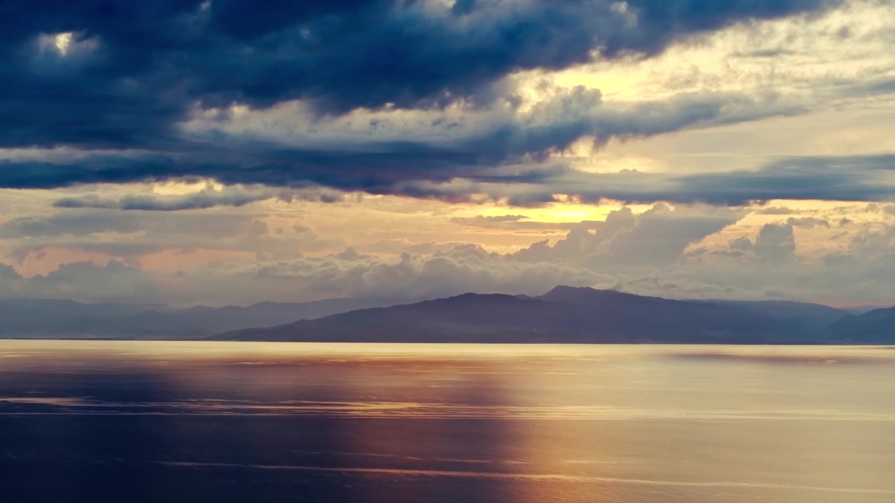 Dark blue storm clouds contrast warm sunset glow over calm sea and distant hills