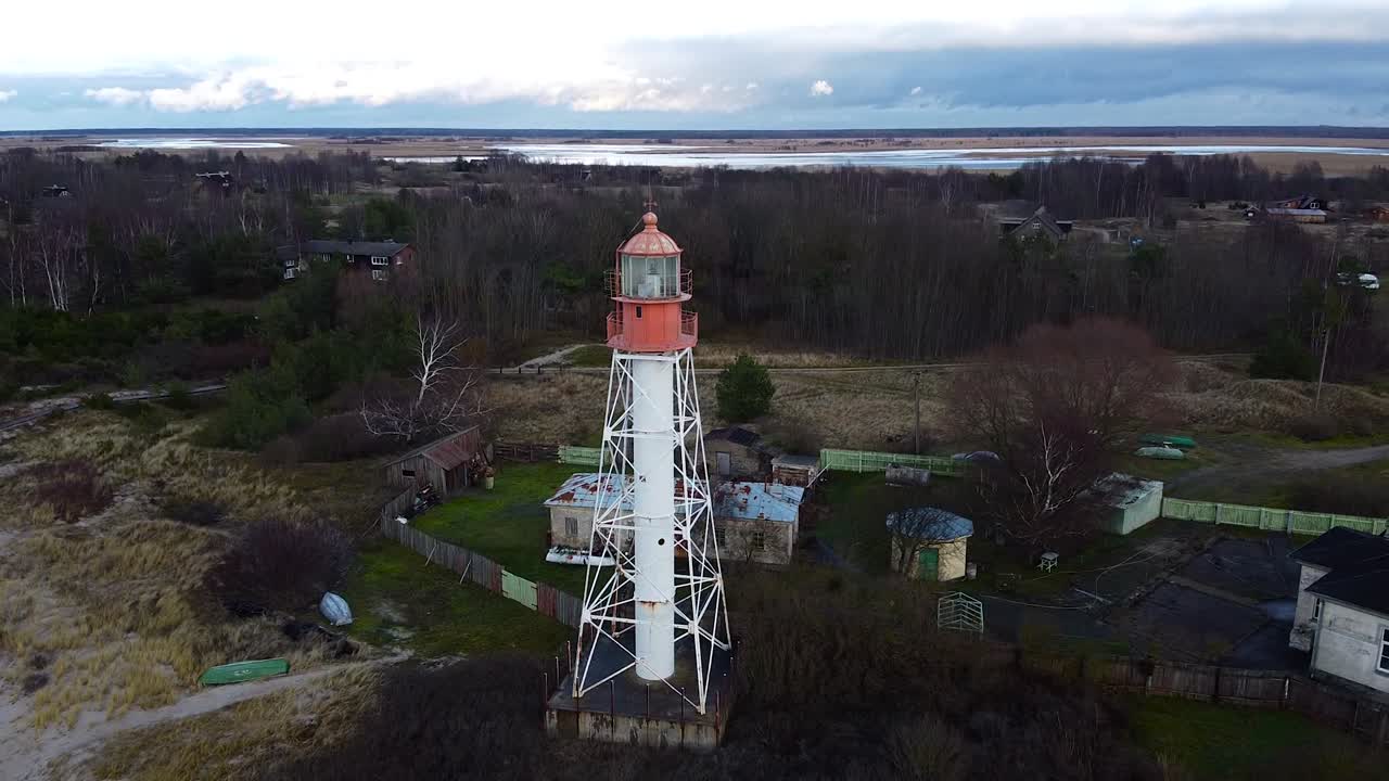 Aerial view of white painted steel lighthouse located in Pape, Latvia at Baltic sea coastline in cloudy day, wide angle establishing drone tilt up shot moving backwards