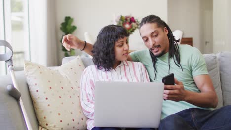 feliz pareja biracial abrazándose en la sala de estar usando computadora portátil y teléfono inteligente, muleta apoyada en el sofá