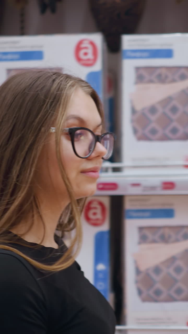 mujer joven caminando más allá de la estantería de la tienda abastecida con productos de cama, ella mira a su alrededor, examinando diferentes opciones de edredones y almohadas, el entorno de la tienda con textiles para el hogar cuidadosamente dispuestos