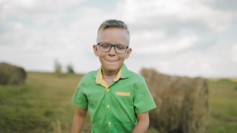 un joven alegre con gafas y una camisa verde con cuello amarillo se encuentra en un vasto campo abierto bajo un cielo brillante, una mujer con un traje casual está borrosa en el fondo