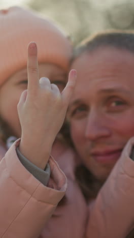 a man wearing a brown jacket carries a little girl in a pink cap and jacket as she makes a playful hand sign with both hands and they smile joyfully