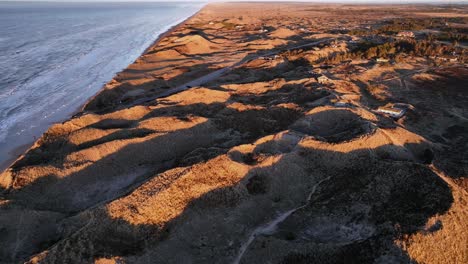 vista a vista de pájaro, grandes dunas de arena de pie junto al mar iluminadas por la luz del atardecer, una casa de verano se encuentra en las dunas, y el agua del mar lava la costa