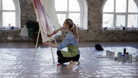 Artist-working-with-large-paintbrush-and-wide-canvas-in-workshop-standing-on-knees