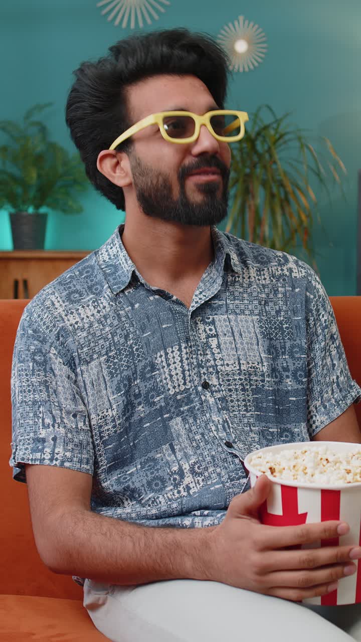 Uomo indiano seduto sul divano che mangia popcorn e guarda un'interessante serie TV o una partita sportiva online a casa.