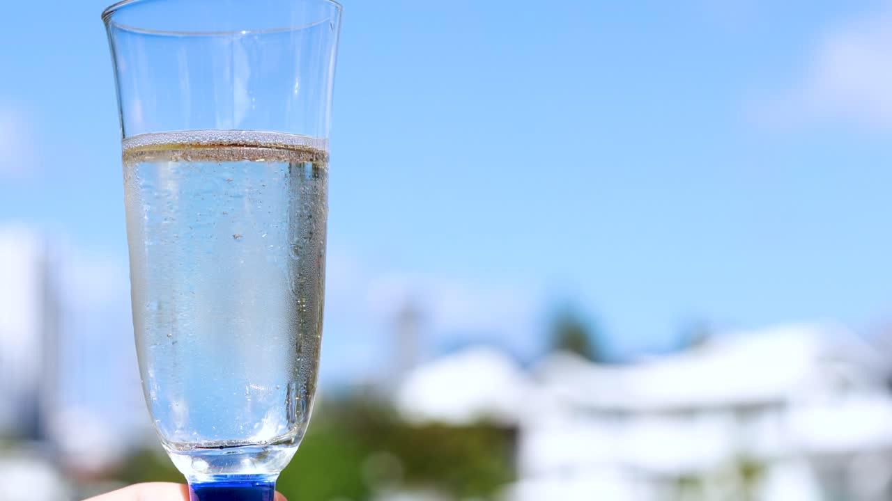 A hand holds a champagne glass filled with sparkling wine against a bright blue sky and blurred urban background, captured in steady daylight