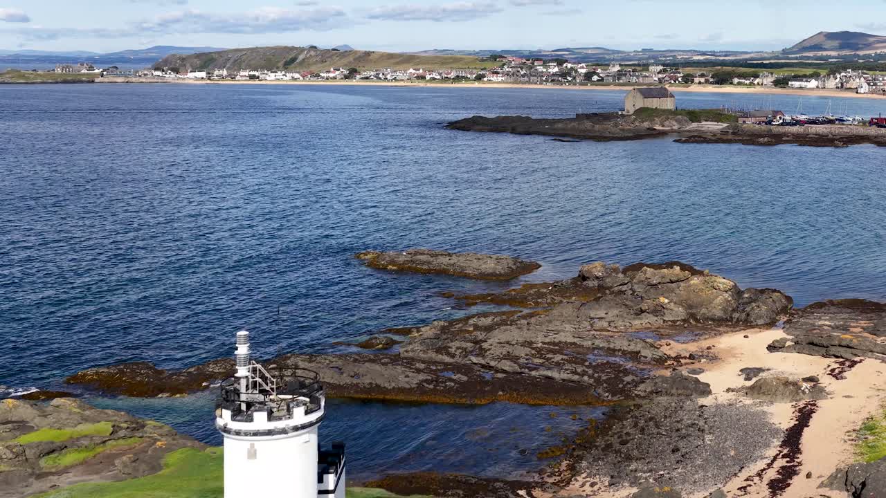Drone pans over rocky shoreline, lighthouse, tents, and seaside village under bright daylight in Scotland