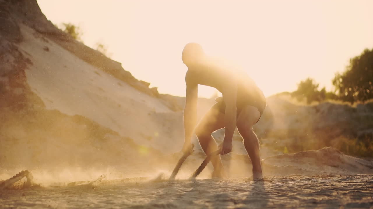 Powerful crossfit training a man with an inflated torso in a buckle hits the ropes powerfully on the ground raising dust and sunlight