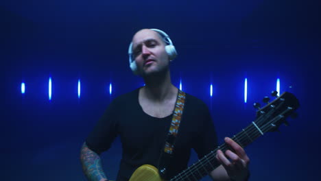 happy male musician playing guitar against bright lights positive tattooed guy in headphones playing guitar against colorful vivid illumination during performance in dark room