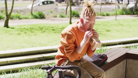 albino african american man with dreadlocks sitting in park with bike eating sandwich