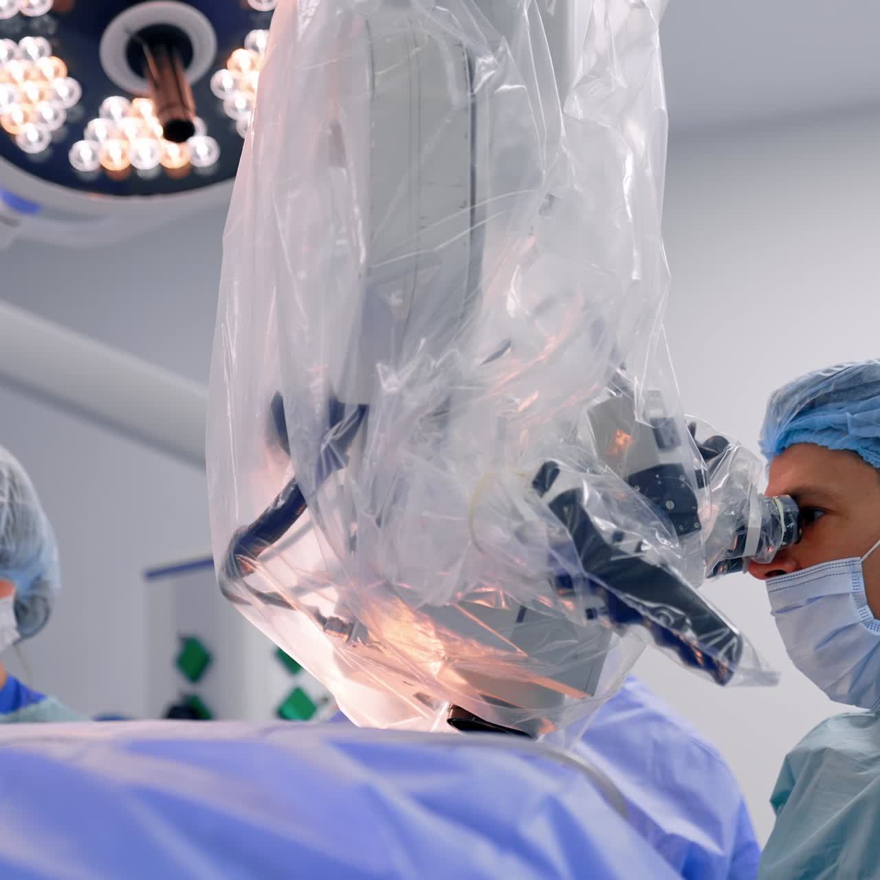 Giovani neurochirurghi seduti che guardano un potente microscopio coperto di plastica. Infermiera sullo sfondo che assiste.