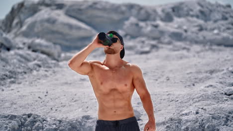 bearded athlete with black flask in nature. shirtless muscular bodybuilder drinking water on white rocky background in summertime.