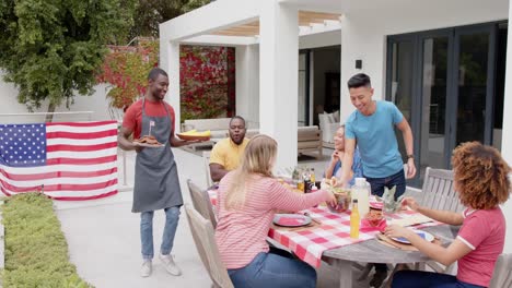 african american man serving food to diverse group of friends at dinner table in garden, slow motion