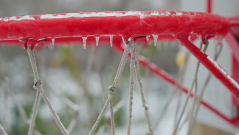 borde de baloncesto rojo vivo cubierto de hielo con hielo colgante, primer plano, contra un patio de recreo nevado borroso, ilustrando el efecto del invierno en las instalaciones deportivas al aire libre