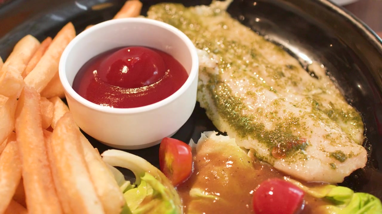 Primo piano di pesce alla griglia servito con patatine fritte croccanti, pesto e ketchup su piatto nero.