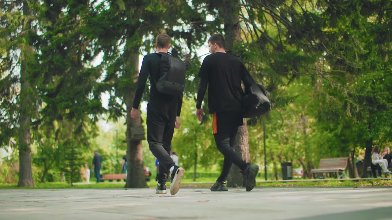 two white men walking through park, backpacks and duffel, casual black clothing, slow reflective conversation, tree lined path with benches and dappled sunlight, relaxed afternoon mood