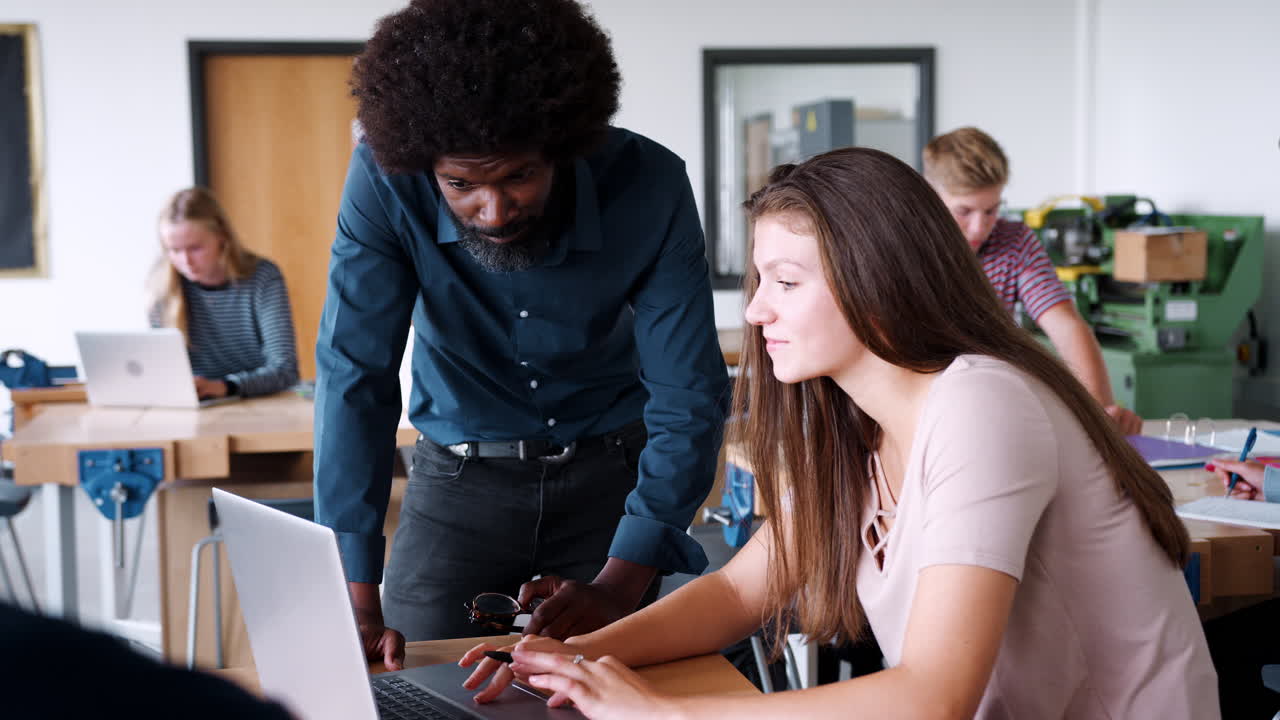 Teacher Talking To Female High School Student Sitting At Work Bench Using Laptop In Design And Technology Lesson
